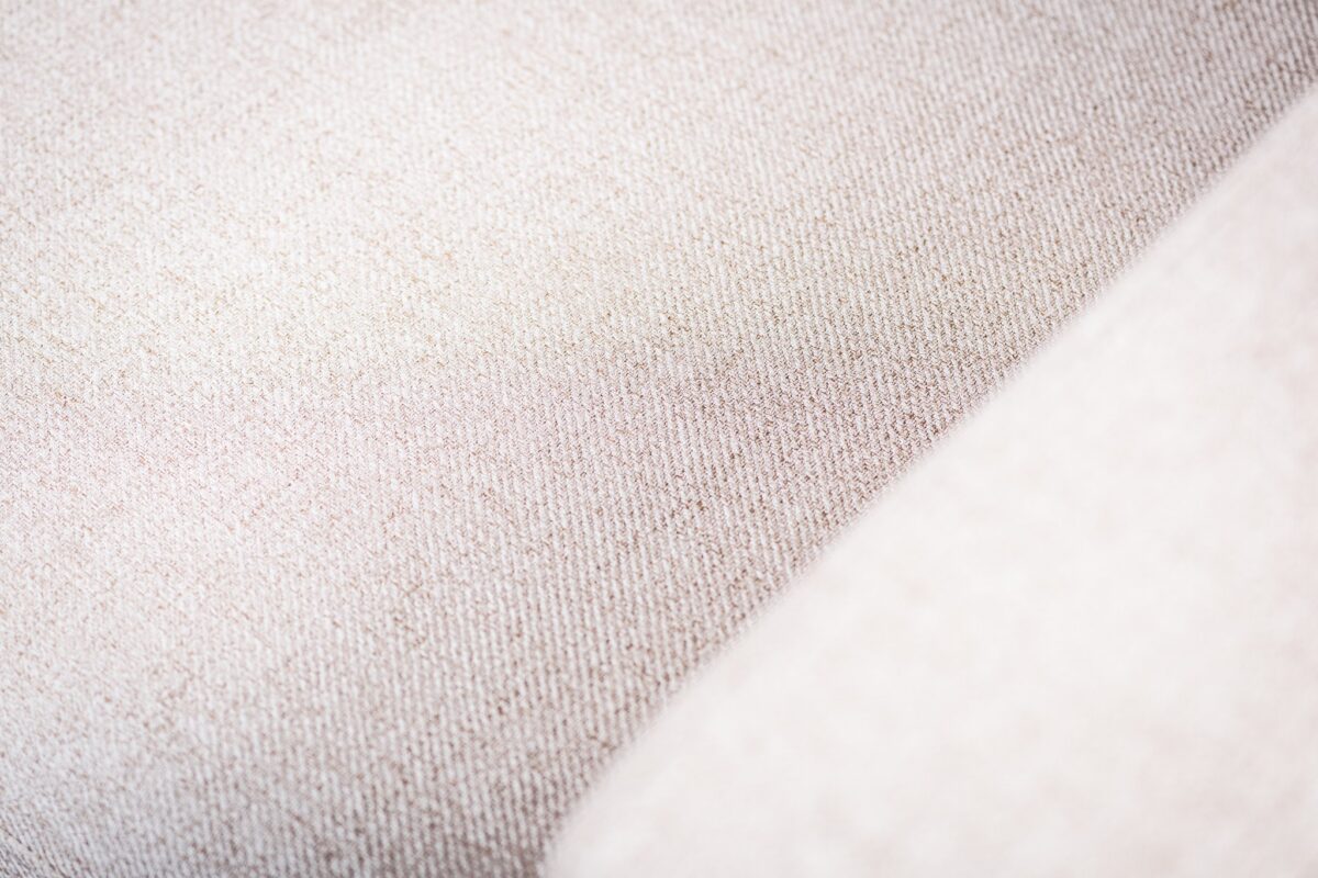 Close-up van een beige stoffen oppervlak met een subtiele geweven textuur. De belichting toont lichte variaties in de kleur van de stof, wat een zachte en neutrale uitstraling creëert.