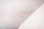 Close-up van een beige stoffen oppervlak met een subtiele geweven textuur. De belichting toont lichte variaties in de kleur van de stof, wat een zachte en neutrale uitstraling creëert.