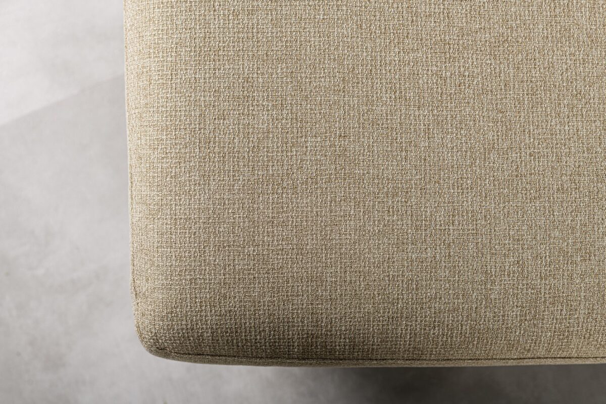 Close-up van een getextureerd beige stoffen oppervlak, mogelijk een gestoffeerd meubelstuk, tegen een lichtgrijze achtergrond. De stof heeft een geweven uiterlijk, wat een eenvoudige en minimalistische esthetiek creëert.