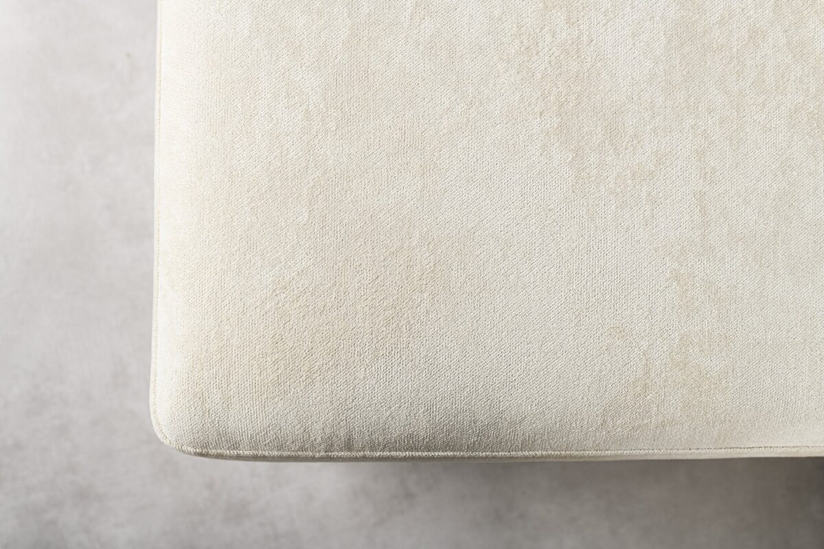 Close-up van een lichtbeige stoffen textuur, met een hoek van een gewatteerd object, mogelijk een bank of stoel, gepositioneerd in de linkerbovenhoek. De achtergrond is een licht getextureerd, lichtgrijs oppervlak.