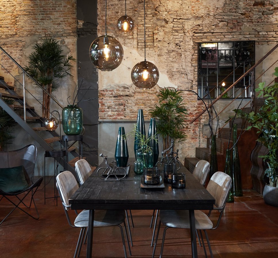 Eetgedeelte in industriële stijl met bakstenen muren, een donkere houten tafel en metalen stoelen. Hangende glazen hanglampen verlichten de ruimte. Groene planten en decoratieve vazen zorgen voor contrast met de rustieke setting. Trappen zijn zichtbaar op de achtergrond.