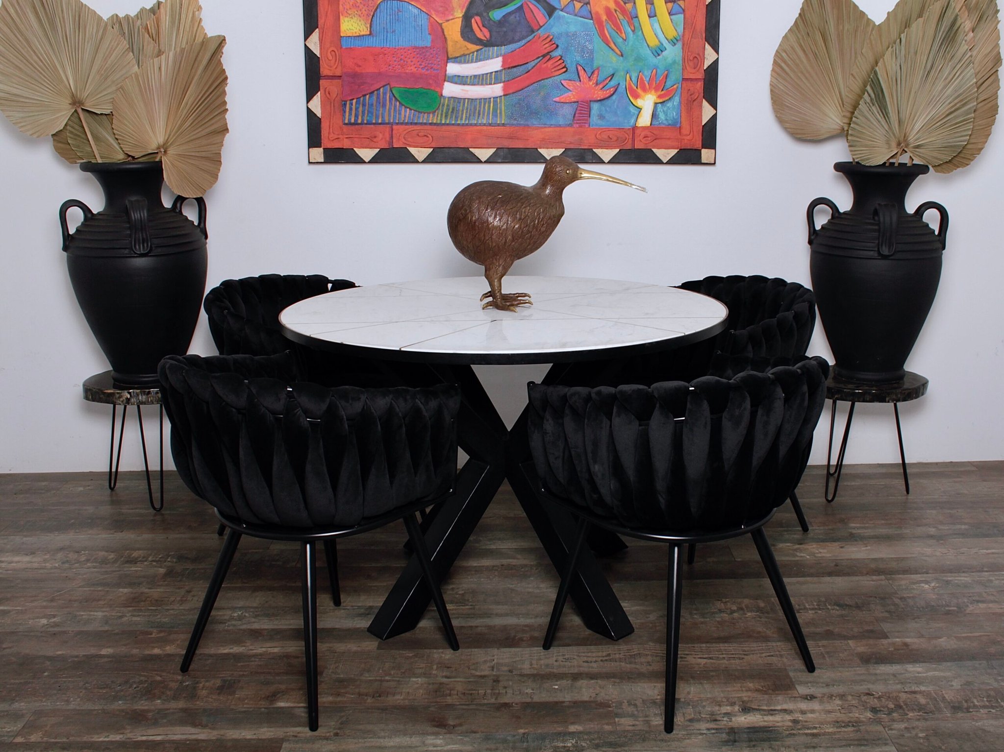 Een eetkamer heeft een ronde marmeren tafel met een kiwi-vogelsculptuur als middelpunt. Rondom de tafel staan zes zwarte gestoffeerde stoelen. Grote urnen met gedroogde palmbladeren flankeren een kleurrijk abstract kunstwerk aan de muur. De vloer is van donker hout.