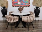 Een ronde eettafel met een wit marmeren blad wordt omringd door zes beige, gecapitonneerde stoelen. Op de tafel staat een kiwi-vogelsculptuur. Aan weerszijden staan twee zwarte urnen met gedroogde palmbladeren. Aan de witte muur hangt een kleurrijk, abstract schilderij.