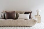 Een gezellige beige bank met zachte kussens in tinten bruin, grijs en crème. Een ronde bijzettafel met een moderne lamp en een kleine plant ernaast. De kamer heeft een minimalistische en serene ambiance met een pluchen wit kleed op de vloer.