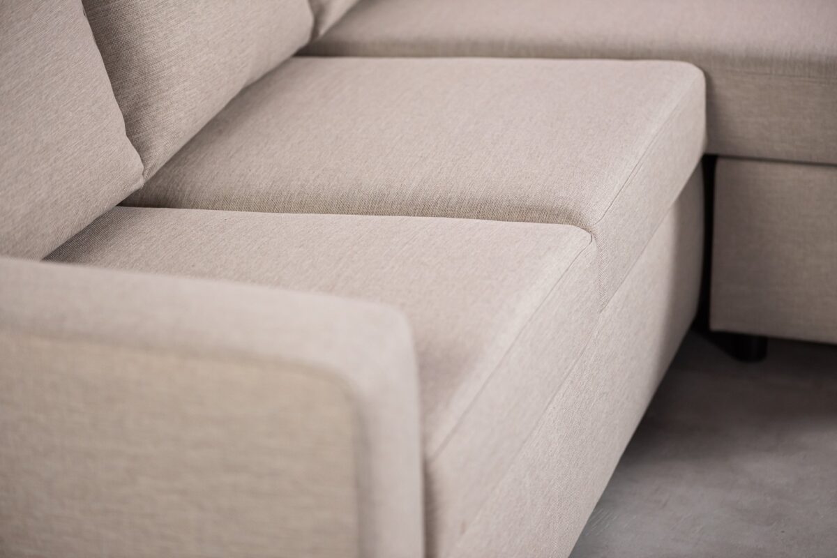 Close-up van een lichtbeige sectionele bank op een grijze vloer. De afbeelding richt zich op de hoek waar twee secties elkaar ontmoeten, waardoor de gladde, zachte stoffen bekleding en de zachte zitting worden benadrukt.