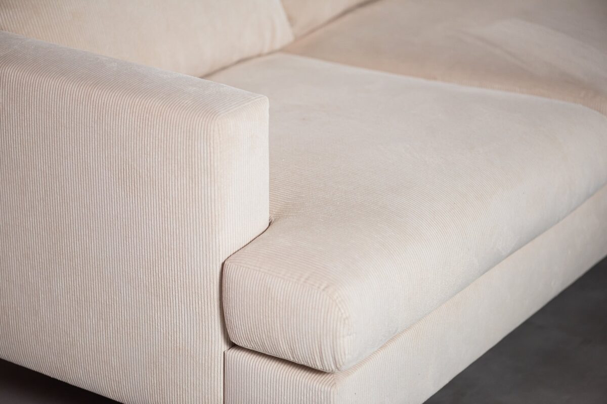 Close-up van een beige bank met een corduroy-textuur, met een zichtbare armleuning en kussen. De stof heeft een subtiel strepenpatroon en de bank staat op een gladde, donkere vloer.