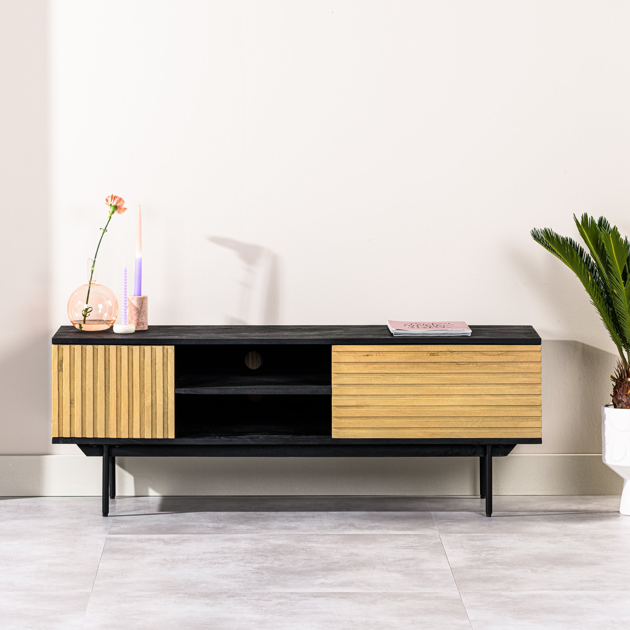 Een moderne houten tv-standaard met zwarte accenten en schuifdeuren. Een roze vaas met een bloem, een kaars en een boek sieren de bovenkant. Een kleine potplant staat ernaast op een lichtgrijze betegelde vloer.