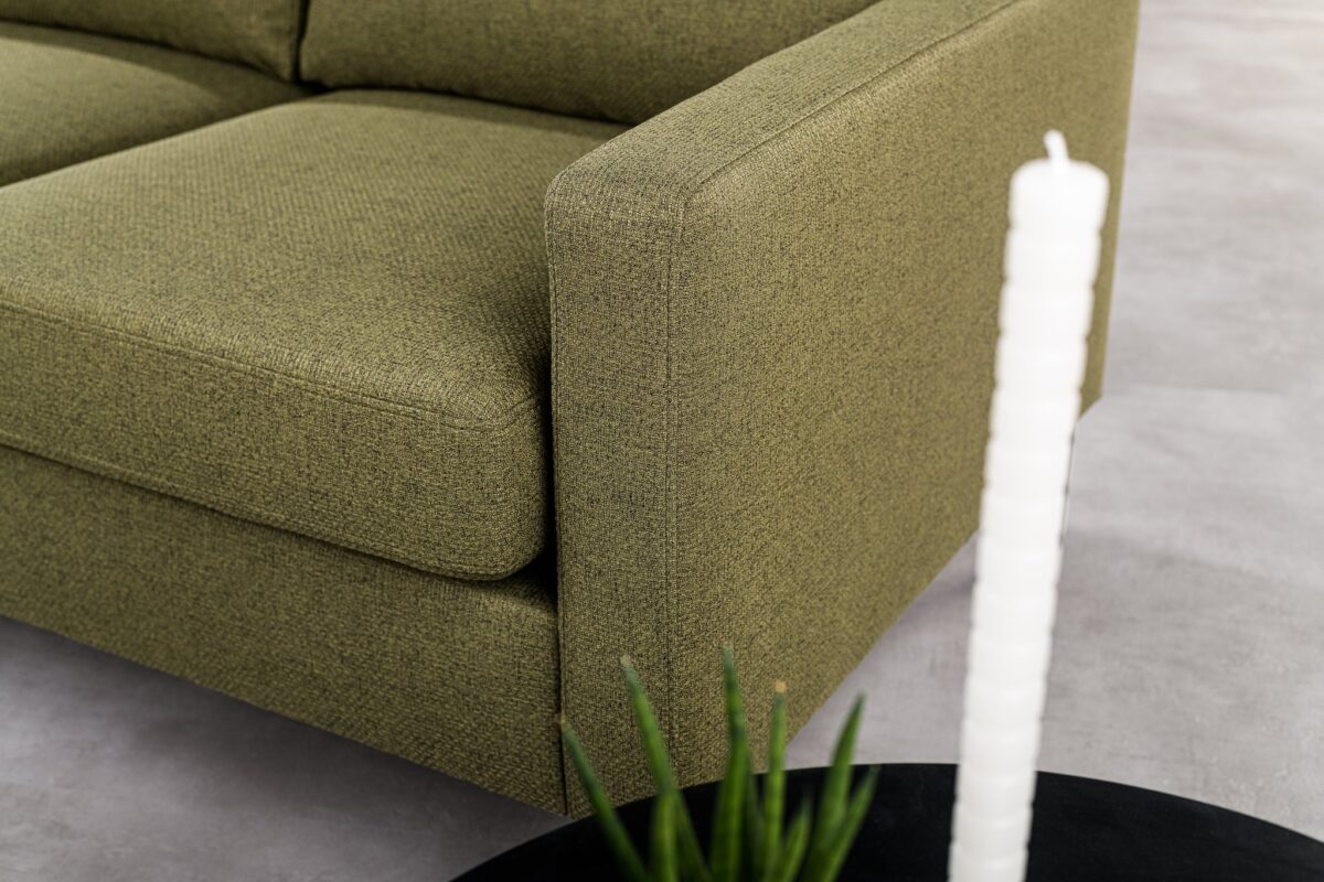 Een close-up van de 3 zits bank CL L+R in Dillon D156 groene stof, met vierkante armleuningen, naast een ronde zwarte tafel met een kaars met witte textuur en een klein groen plantje op een lichtgrijze vloer.