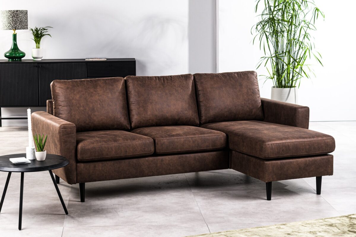 Een bruine leren hoekbank met een L-vormig ontwerp staat in een moderne woonkamer. Een kleine ronde zwarte tafel met een plant en kaars staat ervoor. Op de achtergrond zijn een zwarte console en een hoge groene plant zichtbaar.