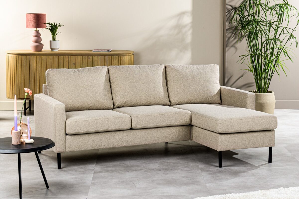 Een beige hoekbank staat in een moderne woonkamer met grijze vloer. Een zwarte ronde bijzettafel met een vaas staat links. Een houten console met decoratieve items en een hoge groene plant staan op de achtergrond tegen een lichtgekleurde muur.