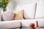 Een beige bank met een rond kussen met textuur en een gestreept rechthoekig kussen. Een vaas met kleurrijke bloemen staat op de achtergrond, wat een vleugje kleur en levendigheid toevoegt aan de serene, neutrale setting.