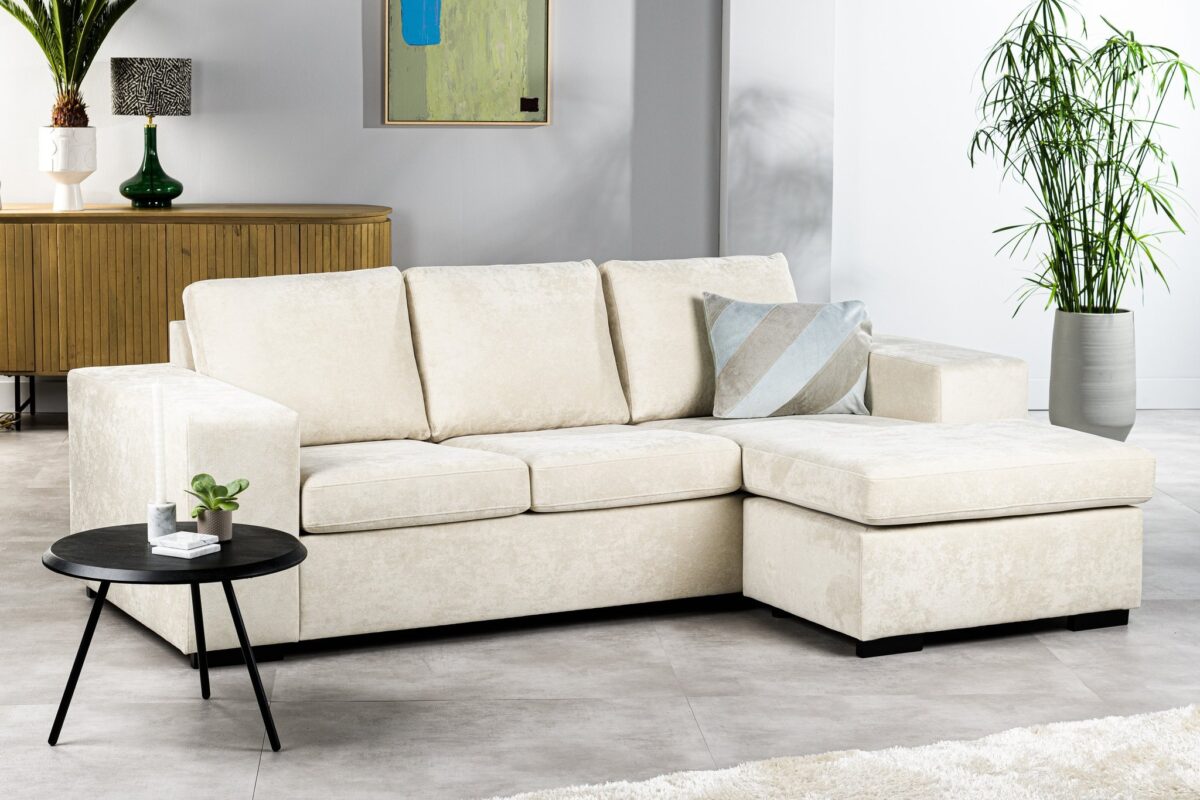 Moderne woonkamer met een beige L-vormige bank, met een geometrisch kussen. Een kleine ronde zwarte tafel met een potplant op de voorgrond. Een houten dressoir, kunstwerken en een hoge plant sieren de achtergrond.