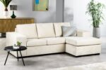 Moderne woonkamer met een beige L-vormige bank, met een geometrisch kussen. Een kleine ronde zwarte tafel met een potplant op de voorgrond. Een houten dressoir, kunstwerken en een hoge plant sieren de achtergrond.