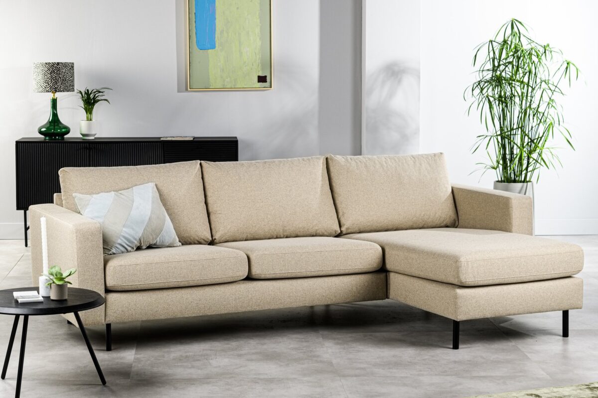 Een moderne woonkamer met een beige hoekbank en een gestreept kussen, naast een kleine ronde tafel met een plant. Op de achtergrond staat een zwarte kast met een lamp en een plant, met daarbovenop een ingelijst kunstwerk. Een hoge potplant staat ernaast.