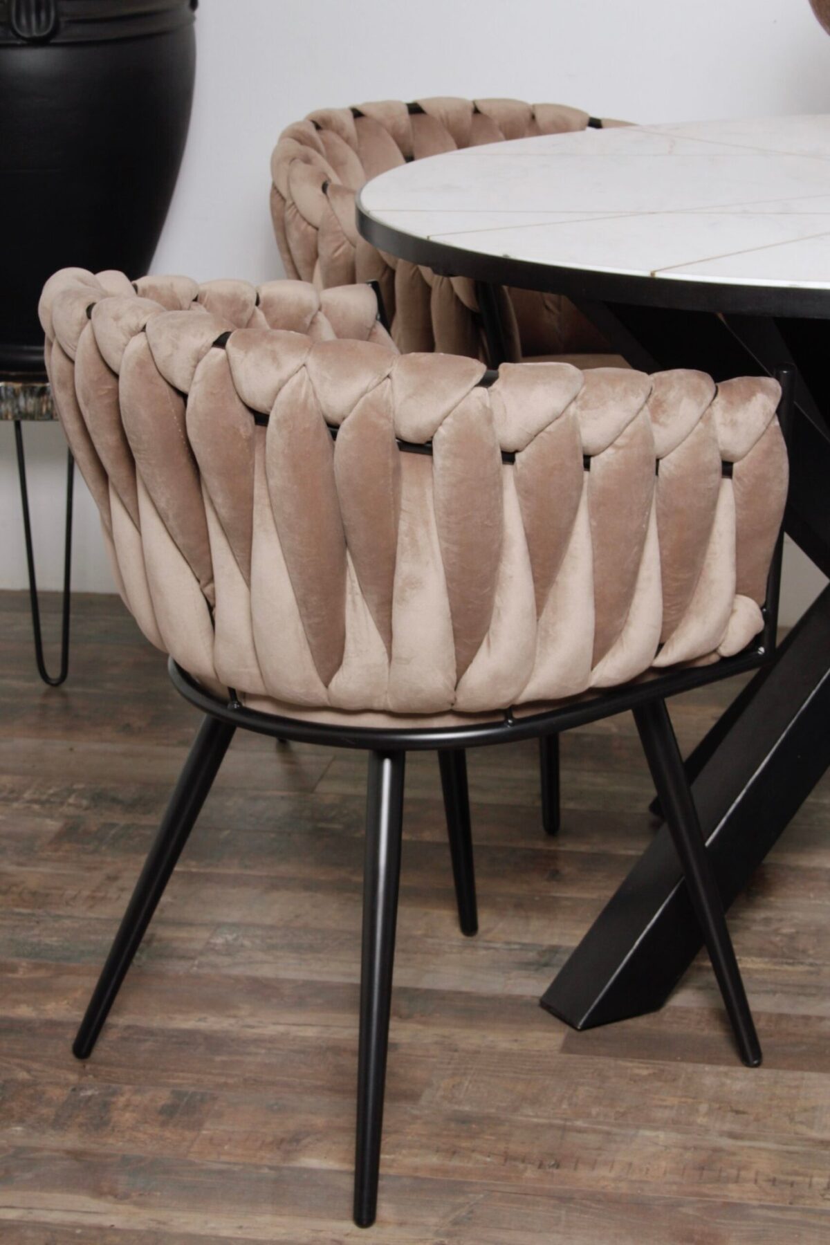 Een bruine, fluwelen stoel met een geweven patroon op de rugleuning staat naast een ronde tafel met een wit blad. De stoel heeft zwarte metalen poten en staat op een houten vloer.