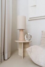 Een minimalistische hoek heeft een beige cilindrische lamp op een klein houten bijzettafeltje. Ernaast staat een modern zilveren beeldhouwwerk. Zachte beige gordijnen en een kussen met textuur versterken het neutrale palet.