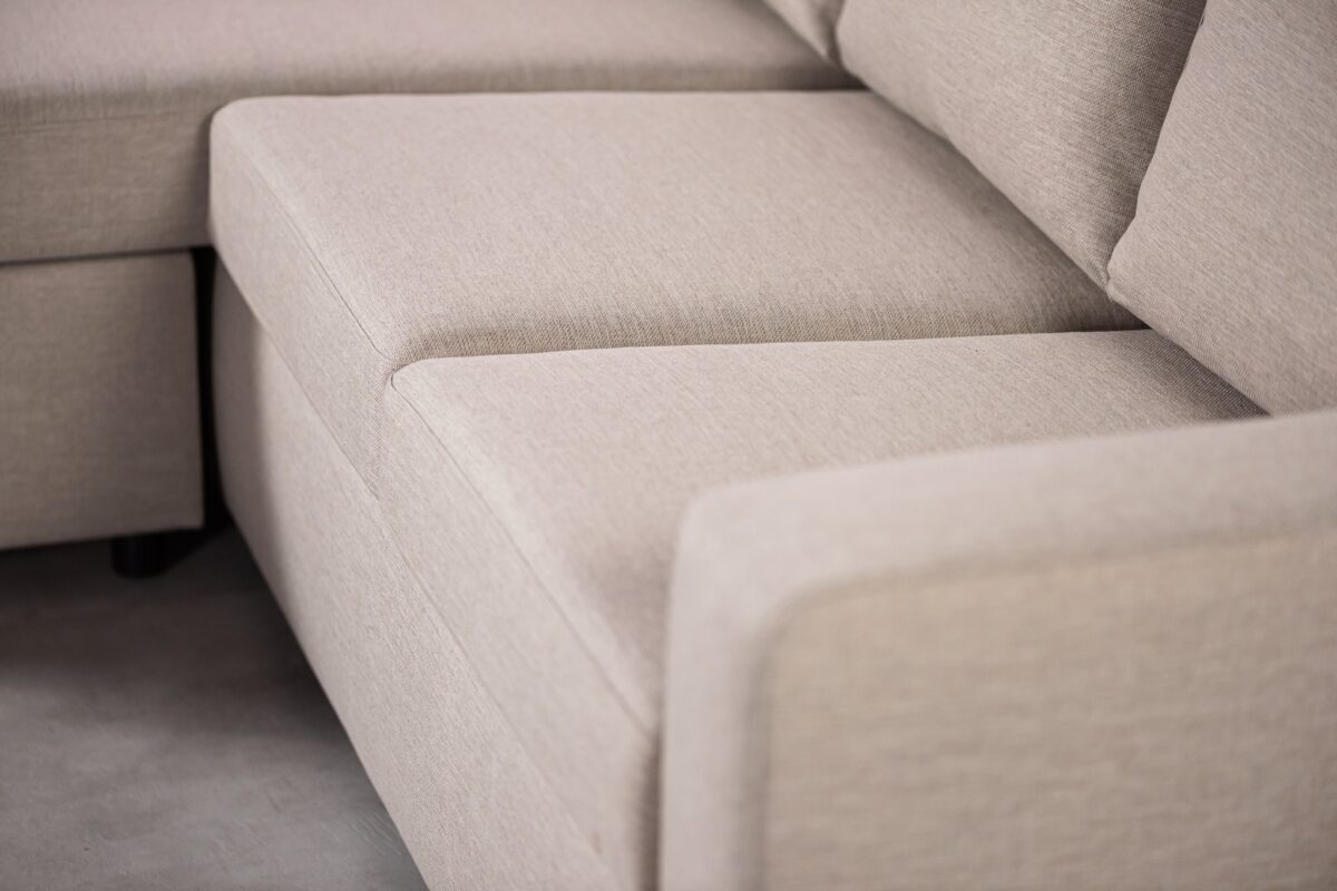 Close-up van een beige sectionele bank met strakke lijnen en zachte kussens, die een modern, minimalistisch design laat zien. De stof heeft een subtiele textuur en de armleuning van de bank is zichtbaar op de voorgrond.