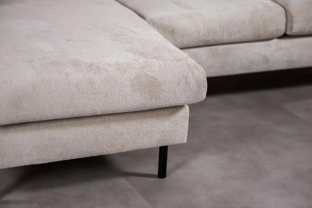 Close-up van een beige sectionele bank met een zachte, gestructureerde stof. De bank heeft strakke, minimalistische lijnen en rust op slanke zwarte metalen poten. Hij staat op een lichtgrijze, gladde vloer.
