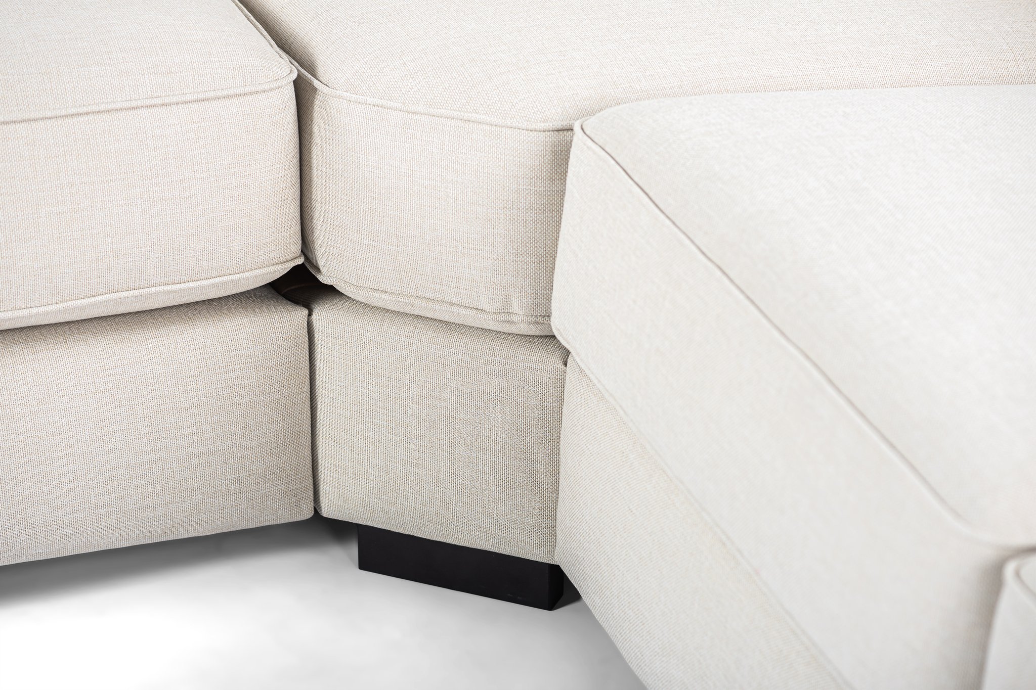 Close-up van een beige, getextureerde sectionele bank met gestoffeerde kussens. De afbeelding richt zich op de hoek waar twee secties elkaar ontmoeten, met daaronder een zwarte bankpoot. De stof heeft een subtiel, geweven patroon, wat bijdraagt aan het moderne en minimalistische ontwerp.