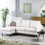Een moderne woonkamer met een witte L-vormige bank en een rond kussen. Er staat een ronde zwarte bijzettafel met een plant en een kaars. Een grote potplant staat ernaast, met een houten dressoir en abstracte kunst aan de muur op de achtergrond.