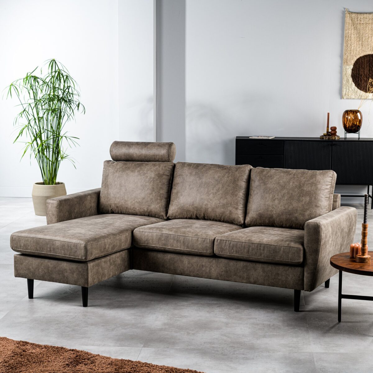 Een grijze sectionele bank met een chaise longue aan de linkerkant staat in een moderne woonkamer. De kamer heeft een hoge potplant, een donker dressoir, decoratieve items en een geweven wandkleed. Een kleine ronde houten tafel staat naast de bank op een lichte tegelvloer.