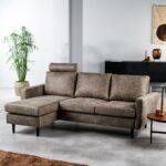 Een grijze sectionele bank met een chaise longue aan de linkerkant staat in een moderne woonkamer. De kamer heeft een hoge potplant, een donker dressoir, decoratieve items en een geweven wandkleed. Een kleine ronde houten tafel staat naast de bank op een lichte tegelvloer.