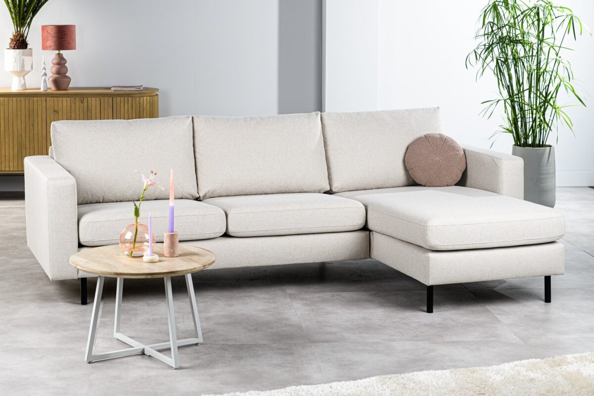 Een moderne woonkamer met de 3 zits bank CL L+R in Dillon D820 ivoor, een ronde houten salontafel met kaarsen en bloemen, een hoge groene plant en een houten dressoir. De ruimte voelt minimalistisch en luchtig aan.