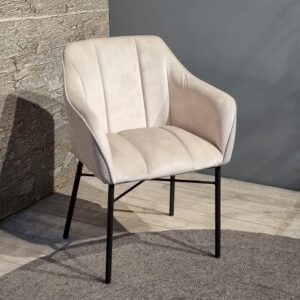De Eetkamerstoel Malaga, een moderne lichtbeige gestoffeerde stoel met zwarte metalen poten, staat op een houten vloer naast getextureerde stenen en grijze muren. Een rond grijs vloerkleed steekt onder de stoel uit.