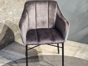 De Eetkamerstoel Malaga, een moderne, grijze, fluwelen accentstoel met zwarte metalen poten, staat op een rond kleed op een lichte houten vloer, naast een muur van getextureerde steen en een gladde, grijze kleur.