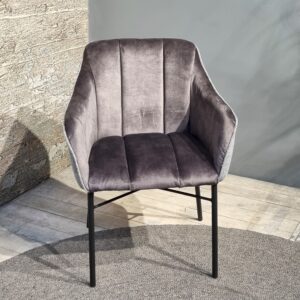 De Eetkamerstoel Malaga, een moderne, grijze, fluwelen accentstoel met zwarte metalen poten, staat op een rond kleed op een lichte houten vloer, naast een muur van getextureerde steen en een gladde, grijze kleur.