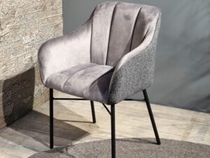 De Eetkamerstoel Malaga heeft een zachte, grijze bekleding, licht gebogen armleuningen, verticale stiksels in de rugleuning en zwarte metalen poten. De stoel staat op een houten vloer naast een muur met structuur.