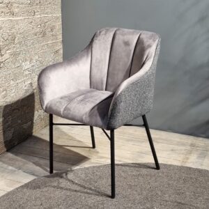 De Eetkamerstoel Malaga heeft een zachte, grijze bekleding, licht gebogen armleuningen, verticale stiksels in de rugleuning en zwarte metalen poten. De stoel staat op een houten vloer naast een muur met structuur.