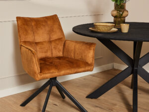Moderne eethoek met de Eetkamerstoel Dani | Velvet Gold, een ronde zwarte tafel met een X-vormig onderstel en een vaas met kleurrijke bloemen, alles op een lichte houten vloer tegen een beige muur.