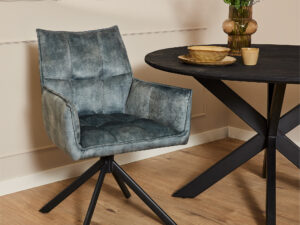 De Eetkamerstoel Dani | Velvet Light Blue, met een strakke fluwelen bekleding en zwarte metalen poten, staat naast een ronde zwarte tafel met een houten blad. Een vaas met kleurrijke bloemen, een kom en een kopje staan op tafel in de lichte kamer.