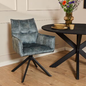De Eetkamerstoel Dani | Velvet Light Blue, met een strakke fluwelen bekleding en zwarte metalen poten, staat naast een ronde zwarte tafel met een houten blad. Een vaas met kleurrijke bloemen, een kom en een kopje staan op tafel in de lichte kamer.