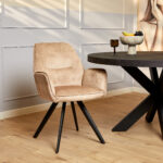 De Eetkamerstoel Nova | Beige, met zwarte schuine poten, staat naast een ronde donkere tafel in een lichte kamer met een lichte houten vloer en witte lambrisering. Een fruitschaal en kunstwerken maken de achtergrond compleet.