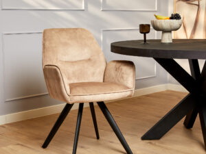 De Eetkamerstoel Nova | Beige, met zwarte schuine poten, staat naast een ronde donkere tafel in een lichte kamer met een lichte houten vloer en witte lambrisering. Een fruitschaal en kunstwerken maken de achtergrond compleet.