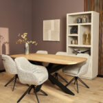Moderne eetkamer met een lichte houten tafel, vijf Eetkamerstoel Jens | Beige stoelen, een hoge witte boekenkast met decoratie, geometrische wandkunst en een vaas met gouden takken op tafel.