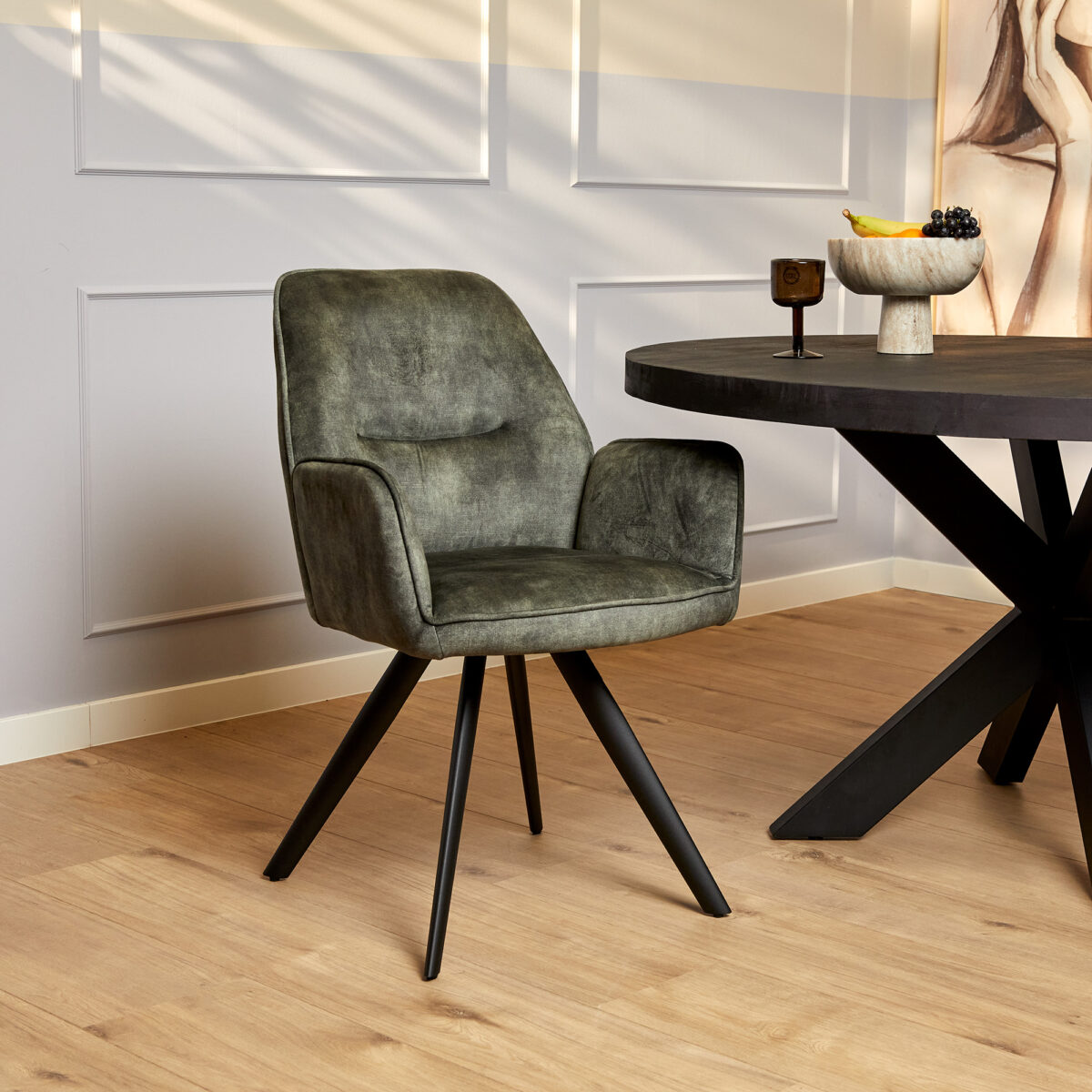 De Eetkamerstoel Nova | Hunter Green, met zwarte schuine poten, staat naast een ronde zwarte eettafel met een fruitschaal en decoratie. De inrichting heeft een lichte houten vloer en lichtgekleurde lambrisering.