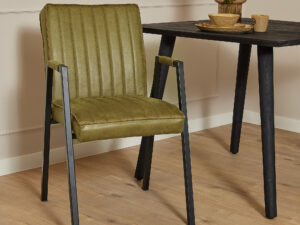 De Eetkamerstoel Tivoli met armleuning in mosgroene stoffering met zwarte metalen poten staat naast een zwarte tafel met lichtbruin keramisch servies en bloemen, het geheel op een lichte houten vloer in een moderne, minimalistische kamer.