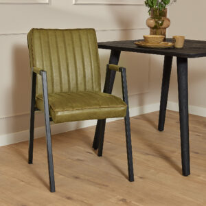 De Eetkamerstoel Tivoli met armleuning in mosgroene stoffering met zwarte metalen poten staat naast een zwarte tafel met lichtbruin keramisch servies en bloemen, het geheel op een lichte houten vloer in een moderne, minimalistische kamer.