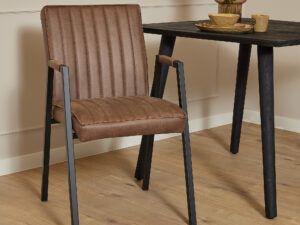 De Eetkamerstoel Tivoli met armleuning | Bull Brown met zwarte metalen poten staat naast een zwarte tafel met daarop een vaas, kom en mok op een lichte houten vloer in een moderne, minimalistische kamer.