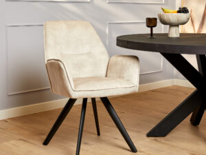 De Eetkamerstoel Nova | Champagne, een stijlvolle beige stoel met zwarte poten, staat naast een ronde zwarte tafel op een houten vloer, tegen een lichtgrijze lambrisering met een fruitschaal als decoratie.