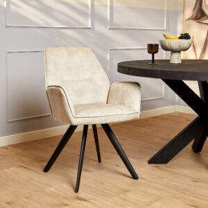 De Eetkamerstoel Nova | Champagne, een stijlvolle beige stoel met zwarte poten, staat naast een ronde zwarte tafel op een houten vloer, tegen een lichtgrijze lambrisering met een fruitschaal als decoratie.