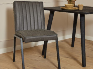 De Eetkamerstoel Tivoli | Bull Antraciet, met grijze bekleding en zwarte metalen poten, staat naast een zwarte houten tafel met servies en een vaas op een lichte houten vloer in een minimalistische kamer.