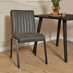 De Eetkamerstoel Tivoli | Bull Antraciet, met grijze bekleding en zwarte metalen poten, staat naast een zwarte houten tafel met servies en een vaas op een lichte houten vloer in een minimalistische kamer.