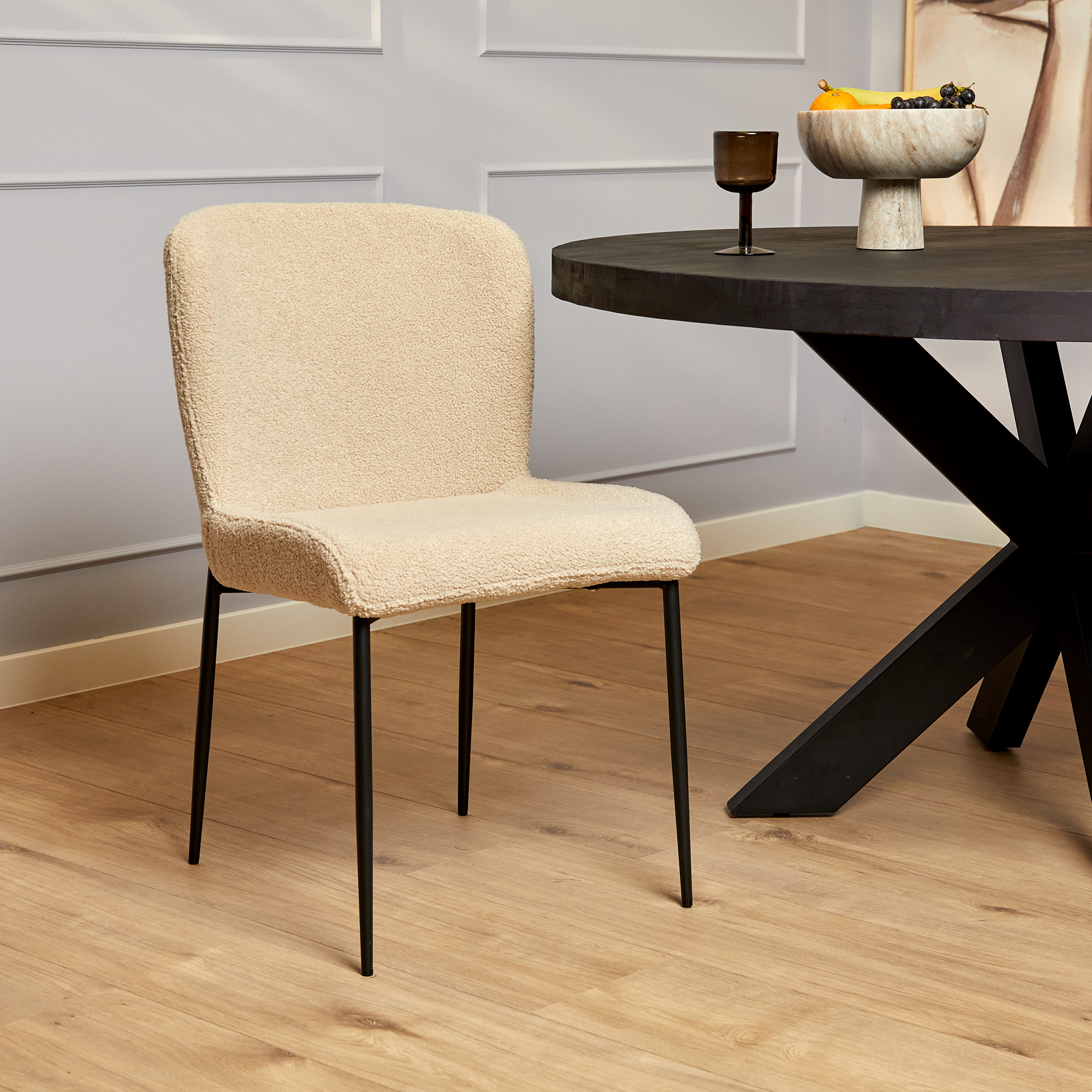 In de moderne eetkamer staat de Eetkamerstoel Tess | Beige met zwarte metalen poten, naast een ronde zwarte tafel met fruit en een donker glas, op een lichte houten vloer en met lichtgekleurde muren.
