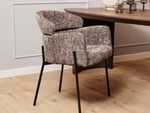 De Eetkamerstoel Otis | Grijs, met structuurgrijze bekleding en zwarte metalen poten, staat naast een donkere houten eettafel op een lichte houten vloer in een kamer met witte lambrisering en decoratieve accenten op de tafel.