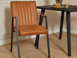De Eetkamerstoel Tivoli met armleuning | Bull Cognac, een bruine leren stoel met zwarte metalen poten, staat naast een zwarte tafel met servies en een vaas met bloemen op een lichte houten vloer tegen een beige muur met lijstwerk.