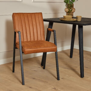 De Eetkamerstoel Tivoli met armleuning | Bull Cognac, een bruine leren stoel met zwarte metalen poten, staat naast een zwarte tafel met servies en een vaas met bloemen op een lichte houten vloer tegen een beige muur met lijstwerk.
